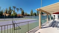 This outdoor tennis facility comprises multiple full-sized courts finished in a classic green and purple hard-court surface, clearly lined and well-maintained for competitive or recreational play. The courts are fully enclosed by green chain-link fencing fitted with privacy windscreen fabric, providing both visual screening and wind protection during play. Tall light poles are positioned throughout the complex, allowing for evening use after sundown.

Entry to the facility is marked by a wooden pergola structure, offering a shaded transition point before accessing the courts. A stucco-clad support building sits adjacent to the courts, featuring green-painted doors — one solid panel door and one multi-pane glass door — providing access to what appears to be storage or facility support rooms. The building's exterior reflects a Spanish-influenced architectural style with a tiled roofline consistent with the surrounding community structures visible in the background.

The facility is set within a lushly landscaped environment, with tall palm trees and mature pines framing the perimeter, creating a resort-like atmosphere. Surrounding architecture, including red-tile-roofed buildings, reinforces the cohesive aesthetic of the broader community. The entire complex benefits from abundant natural sunlight under clear skies, making it an inviting and functional space for tennis enthusiasts.