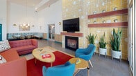Located at 3530 E La Palma Ave in Anaheim, California, this single-story community clubhouse showcases a bold mid-century modern aesthetic with a lively, curated design throughout. The main lounge area commands attention with soaring double-height ceilings and a dramatic statement fireplace clad in patterned yellow tile, framed by terracotta-toned shelving and a mounted flat-screen television. Vibrant furnishings — including a coral sofa, teal accent chairs, and a red area rug — bring energy and character to the space. Floor-to-ceiling glass windows and sliding doors flood the interior with abundant natural light while offering unobstructed views of lush palm trees and an outdoor patio area, seamlessly connecting indoor and outdoor living. A cozy dining nook features plaid-upholstered wraparound banquette seating, artful wall hangings, and a striking multi-globe pendant chandelier that adds a retro-modern touch. The full community kitchen is equipped with a large center island, built-in appliances, sleek cabinetry in light wood tones, and a vibrant turquoise accent wall, all illuminated by a playful array of colorful cone pendant lights. Large-format tile flooring flows throughout, unifying the space with a clean, contemporary finish.
