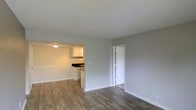 This single-floor space offers a clean, functional layout designed for comfortable everyday living. The main living area is finished with wood-look plank flooring that adds warmth and visual continuity throughout the space. Neutral gray walls and white trim create a fresh, modern backdrop that suits a variety of interior styles.

The kitchen is accessible directly from the living area through a half-wall pass-through opening, maintaining an open and connected feel. It features white upper cabinetry, a dark countertop, and an under-cabinet light fixture that provides focused task lighting. A sink is positioned along the kitchen wall, and the space is designed for efficient use.

A doorway visible from the main living area leads to an additional room, suggesting a private space such as a bedroom or storage area, complete with its own door for privacy. The textured popcorn ceiling adds a classic residential touch, while flush-mounted lighting provides ambient illumination throughout.

Overall, the space presents a straightforward and practical layout with cohesive finishes, offering a well-lit and neatly maintained environment that balances open communal areas with defined private spaces.