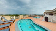 Located atop a prominent building in Athens, Georgia, this rooftop pool deck offers an exceptional outdoor retreat with commanding views of the surrounding cityscape and treeline. The centerpiece of the space is a generously sized, curved swimming pool with a vivid turquoise finish, complemented by a separate circular hot tub positioned nearby for easy accessibility. The entire deck is finished with warm terracotta-toned tiles that extend throughout, creating a cohesive and inviting atmosphere. Multiple sets of cushioned lounge chairs are arranged poolside, providing ample space for relaxation while taking in the expansive open-sky views. A built-in wet bar area featuring a granite countertop and sink adds a convenient amenity for poolside entertaining. Decorative white lattice-style perimeter railings frame the deck elegantly while maintaining unobstructed sightlines to the Athens horizon. A stone-clad utility structure on the deck houses support facilities and adds an architectural accent to the rooftop environment. Potted greenery and additional seating areas with table-and-chair sets round out the space, making it suitable for both casual lounging and social gatherings. This rooftop pool deck delivers a well-appointed, elevated outdoor experience in the heart of Athens.