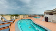 Located atop a prominent building in Athens, Georgia, this rooftop pool deck offers an exceptional outdoor retreat with commanding views of the surrounding cityscape and treeline. The centerpiece of the space is a generously sized, curved swimming pool with a vivid turquoise finish, complemented by a separate circular hot tub positioned nearby for easy accessibility. The entire deck is finished with warm terracotta-toned tiles that extend throughout, creating a cohesive and inviting atmosphere. Multiple sets of cushioned lounge chairs are arranged poolside, providing ample space for relaxation while taking in the expansive open-sky views. A built-in wet bar area featuring a granite countertop and sink adds a convenient amenity for poolside entertaining. Decorative white lattice-style perimeter railings frame the deck elegantly while maintaining unobstructed sightlines to the Athens horizon. A stone-clad utility structure on the deck houses support facilities and adds an architectural accent to the rooftop environment. Potted greenery and additional seating areas with table-and-chair sets round out the space, making it suitable for both casual lounging and social gatherings. This rooftop pool deck delivers a well-appointed, elevated outdoor experience in the heart of Athens.