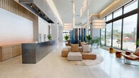 Located in Oakland Park, Florida, this expansive single-floor amenity space offers a sophisticated blend of modern design and functional comfort. The grand lobby serves as the centerpiece, featuring polished concrete flooring, soaring ceilings with elegant pendant lighting, and floor-to-ceiling windows that flood the interior with abundant natural light and offer views of the surrounding Florida landscape. Lush tropical plants are strategically placed throughout, reinforcing the space's connection to its South Florida setting.

The reception area is anchored by a sleek, dark stone front desk, complemented by sculptural pendant fixtures overhead. Multiple seating vignettes are thoughtfully arranged across the open floor plan, including modular lounge furniture in warm neutral tones and unique tiered wooden seating platforms upholstered in bold blue, offering both style and versatility.

Dramatic dark vertical panel accents provide visual contrast against the predominantly white walls and ceilings. A dedicated fitness center, enclosed by full-height glass walls, is fully equipped and benefits from high ceilings and ample natural light. A kitchenette area features warm wood slat wall paneling, built-in cabinetry, and dark countertops. The entry vestibule showcases large bronze-framed glass doors, continuing the space's refined, contemporary architectural language throughout.