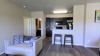 This single-floor unit offers a thoughtfully designed open-concept layout that seamlessly connects the living area to the kitchen. The living space features warm, wood-style flooring that extends throughout, creating a cohesive and inviting atmosphere. A comfortable seating area anchors the living space, accented by neutral tones and natural light.

The kitchen is a standout feature, boasting rich dark cabinetry that contrasts elegantly with light granite countertops. A peninsula with bar-height seating for two provides both additional prep space and a casual dining option. The kitchen is well-equipped with black appliances, including a refrigerator, range, and microwave, making it both functional and stylish.

Just off the main living area, a hallway leads to additional rooms, with a glimpse of a bathroom visible in the background. The bathroom features a well-lit vanity with a dark wood finish, consistent with the overall design aesthetic of the unit.

Neutral-toned walls and recessed ceiling lighting enhance the bright, clean ambiance throughout the space. The consistent use of dark wood tones against light walls and countertops gives the unit a modern, polished look that is both practical and visually appealing.