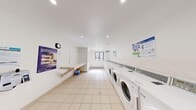 This shared laundry facility is a compact, efficiently organized space designed to serve the practical needs of its users. The room features light-toned ceramic tile flooring throughout, contributing to a clean and hygienic atmosphere. Bright white walls and recessed ceiling lighting create a well-lit, functional environment.

Along one side of the room, a row of front-loading washing machines is positioned adjacent to a matching set of top-loading dryers, providing ample laundry capacity. A built-in wall-mounted folding counter runs along the opposite side, offering a convenient surface for sorting and folding laundry. A payment terminal is mounted near the entrance, enabling easy access to machine operation.

A window on one end of the room allows natural light to filter in, enhancing the brightness of the space. Baseboard heating is present along the walls, ensuring comfort during cooler months. Informational signage is posted throughout, providing usage instructions and contact details for the laundry service provider.

The space is accessed through a secure door and includes a wall clock for user convenience. The overall layout is straightforward and practical, maximizing usability within a modest footprint while maintaining a tidy, well-maintained appearance.