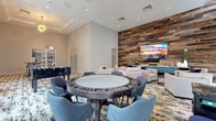 This well-appointed amenity lounge is designed to accommodate both social gatherings and relaxed leisure. The space is anchored by a dramatic floor-to-ceiling reclaimed wood accent wall, against which a large flat-screen television is mounted above a teal-finished media credenza. Arranged before it, a lounge seating area features plush cream armchairs accented with navy throw pillows, centered around a dark wood coffee table and layered over a bold abstract area rug.

The game area occupies a generous portion of the room and includes a dedicated poker/card table surrounded by upholstered blue chairs, as well as a foosball table nearby. High ceilings throughout the space contribute to an open, airy feel, and are complemented by striking gold sputnik chandeliers that add a contemporary touch.

Floor-to-ceiling windows along one wall flood the interior with natural light and offer views of the surrounding greenery. Additional high-top tables with bar-height seating are positioned near the windows, providing casual spots for conversation. Smaller bistro tables are tucked into transitional areas, ensuring the layout accommodates a variety of uses. The overall design blends modern and rustic elements seamlessly.