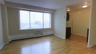 This single-floor unit offers an open, airy layout highlighted by gleaming light hardwood floors that extend throughout the main living area. A large picture window dominates one wall, fitted with vertical blinds and framing expansive views of the surrounding urban skyline, allowing abundant natural light to fill the space. A wall-mounted air conditioning unit sits beneath the window, complementing the baseboard heating system that runs along the perimeter.

Adjacent to the main living area, a compact kitchen is partially separated by dark-toned cabinetry, creating a subtle visual division between the two spaces. The kitchen includes a white refrigerator and is illuminated by a dedicated overhead light fixture. The cabinetry's dark finish provides a striking contrast against the neutral, cream-toned walls throughout the unit.

The overall ambiance is clean and understated, with a minimalist aesthetic defined by light tones, smooth walls, and an absence of clutter. The combination of natural light from the oversized window and the reflective quality of the hardwood floors contributes to a bright, welcoming atmosphere well-suited to urban living.