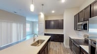 This well-designed single-floor residence offers a thoughtful layout that blends functionality with modern aesthetics. The open-concept kitchen is a standout feature, showcasing rich dark espresso cabinetry with brushed nickel hardware, expansive white quartz countertops, and a full suite of stainless steel appliances including a range, microwave, refrigerator, and dishwasher. A large center island with an undermount sink and elegant pendant lighting serves as both a prep area and a natural gathering point. Generous cabinet and counter space lines the walls, providing ample storage and workspace. The kitchen flows seamlessly into the living area, which is bathed in natural light from multiple windows and features durable wood-look laminate flooring. The bedrooms are carpeted for comfort, with large windows that bring in natural light and offer outdoor views. At least one bedroom includes a walk-in closet, adding practical storage. The primary bathroom is well-appointed with dark cabinetry matching the kitchen, a dual-sink vanity with quartz counters, a glass-enclosed walk-in shower, and wood-look tile flooring. Neutral warm-toned walls and recessed lighting throughout create a clean, cohesive ambiance across the entire space.