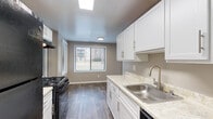 This single-floor residence offers a thoughtfully designed layout with a neutral color palette of warm grays and taupe tones that creates a cohesive, calming atmosphere throughout. The living area is generously sized, featuring plush wall-to-wall carpeting and large vertical-blind-covered sliding glass doors that invite abundant natural light and provide views of the surrounding greenery. The space flows naturally into an open secondary area, defined by an architectural partition, offering flexibility for various living arrangements. The kitchen is a functional and attractive galley-style space finished with wood-look vinyl flooring, crisp white shaker-style cabinetry with brushed nickel hardware, light stone-look countertops, a stainless steel sink, and black appliances including a gas range and refrigerator. A dedicated dining nook with a window completes the kitchen area. The bedrooms are comfortably proportioned, carpeted, and feature ample closet space, including a bifold-door closet in one room, with windows providing natural light and outdoor views. The bathroom is clean and bright, offering a full tub-and-shower combination with white tile surround. Throughout the residence, clean white baseboards and consistent finishes contribute to a polished, move-in-ready presentation.
