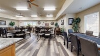 Located at 300 Garden Ln in Weatherford, TX, this well-appointed single-story community facility offers a generously sized, open floor plan designed to serve a variety of communal activities. The main gathering area is expansive and inviting, finished with durable wood-look plank flooring throughout and illuminated by overhead fluorescent lighting complemented by ceiling fans for year-round comfort. This central space accommodates multiple dining and seating arrangements, featuring dark wood tables and chairs, a cozy lounge corner with upholstered sofas, and a dedicated reading nook lined with tall bookshelves stocked with a wide selection of books. Natural light filters in through windows along the perimeter, enhancing the warm, neutral color palette of the walls. The fully equipped kitchen is centrally accessible and showcases a large granite-topped island, light wood cabinetry, black appliances including a refrigerator and range, and tile flooring. A separate exercise room provides a dedicated fitness space outfitted with treadmills, a stationary bike, and a recumbent cycle, all set on the same wood-look flooring and kept comfortable by a ceiling fan. Together, these spaces create a functional and welcoming environment suited for community gatherings, recreation, and daily wellness activities.
