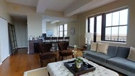This single-floor space showcases an open-concept layout that seamlessly connects a living area with a casual bar or counter seating zone. The living area is anchored by a tufted white ottoman serving as a central coffee table, complemented by richly upholstered dark brown accent chairs with button-tufted detailing. A grey sofa lines one wall, dressed with neutral and cream-toned throw pillows, creating a sophisticated yet comfortable seating arrangement. A decorative floral centerpiece adds a touch of warmth to the space.

The bar area features dark wood counter-height stools with tufted seat backs, positioned along a raised counter. A wine rack and a small collection of bottles are visible, lending a casual entertaining feel. An oversized round wall clock serves as a decorative focal point in this zone.

Structural beam detailing on the ceiling adds architectural character, while multiple windows allow natural light to fill the space. The warm hardwood flooring ties the areas together cohesively. The overall ambiance is polished and inviting, blending classic furnishings with functional entertaining elements.