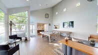 Located at 16201 NW Schendel Ave in Beaverton, Oregon, this two-floor community space offers a sophisticated and welcoming atmosphere designed for both leisure and socializing. The main floor commands attention with its dramatic vaulted ceilings that rise to impressive heights, creating an airy, open ambiance throughout the great room. Expansive floor-to-ceiling windows flood the interior with abundant natural light while framing lush green views of the surrounding trees, seamlessly connecting the indoors with the outdoors. Warm hardwood-style flooring runs throughout, lending a sense of continuity and warmth to the space. The game area is a central highlight, featuring a full-size pool table and a shuffleboard table, both finished in natural wood tones that complement the overall aesthetic. A wall-mounted flat-screen TV and curated artwork, including a standout highland cattle print, add personality to the space. A comfortable lounge area with a sectional sofa, leather seating, and a bold pendant light fixture provides a relaxed retreat. The dining nook, situated on the upper level, offers an intimate setting with plush channel-tufted banquette seating, round marble-topped tables, and a pair of elegant watercolor artworks. A staircase connects the two floors, completing this well-appointed and versatile community gathering space.