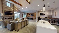 Located at 101 Bill Robinson Way in Newburgh, NY, this single-floor clubhouse is a thoughtfully designed community gathering space that blends rustic warmth with modern sophistication. Soaring vaulted ceilings accented by exposed wooden beams set an inviting tone throughout the open-concept layout, while herringbone-patterned carpeting ties the space together with understated elegance.

The main lounge area serves as the heart of the clubhouse, centered around a dramatic floor-to-ceiling fireplace clad in reclaimed wood tile and topped with a rustic wood mantel. A large flat-screen TV is mounted above the fireplace, with comfortable upholstered seating arranged for relaxed viewing and conversation. French doors along the lounge wall invite natural light and offer views of the outdoor surroundings.

Adjacent to the lounge, a dedicated game zone features a full-size pool table and an arcade game station, providing ample entertainment options. A bar-height counter with industrial-style stools sits against a decorative wall mural featuring a vintage map of the Newburgh region, adding a locally inspired artistic touch.

The kitchenette is outfitted with granite countertops, dark cabinetry, stainless steel appliances including a full-size refrigerator and microwave, and a wall-mounted TV — making it ideal for catered events or casual use. Stylish pendant lighting throughout completes the polished, community-forward atmosphere of this versatile clubhouse.