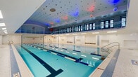 Located on Euclid Avenue in Cleveland, Ohio, this indoor aquatic facility is a striking two-floor space centered around a pristine multi-lane lap pool. The pool is flanked by a generously wide deck finished in intricate mosaic tilework, blending neutral tones with subtle decorative accents — including an emblazoned club logo inlaid directly into the floor. White subway tile lines the perimeter walls, lending a clean, institutional elegance to the space.

The soaring ceiling draws the eye upward, where a vaulted arch crowns one end of the natatorium. Dramatic colored lighting — featuring red and blue accent fixtures — illuminates the ceiling and reflects off the pool's crystal-clear water, creating a vibrant yet sophisticated ambiance. Recessed skylights and flush panel lighting provide consistent, functional illumination along the pool deck.

A full second-floor mezzanine wraps around the pool area, enclosed by a metal railing and lined with large multi-pane windows. This elevated gallery offers an unobstructed view of the pool below and connects via two staircases to the pool level. The upper level's windowed perimeter fills the space with natural light, reinforcing the open, airy character of the facility. Safety equipment, entry doors, and seating areas are thoughtfully integrated throughout the deck level, completing this well-appointed aquatic venue.
