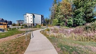 This thoughtfully designed outdoor communal space offers a welcoming environment for residents and visitors alike. A smooth, gently curving concrete pathway winds throughout the grounds, lined with decorative lamp posts that provide both charm and practical illumination. The pathway is flanked by well-kept lawns, native shrubs, ornamental grasses, and mature trees that lend a natural, park-like atmosphere to the setting.

The space includes several distinct amenity areas. A designated picnic zone features a classic wooden picnic table set on a circular concrete pad, ideal for casual outdoor dining or social gatherings. Adjacent natural plantings and shrub beds add texture and visual interest throughout the grounds.

A fenced dog run area is clearly visible, offering a safe and dedicated space for pets to exercise freely. Nearby, a children's playground with timber-framed structures provides recreational opportunities for younger residents.

The surrounding residential buildings are modern in design, featuring bold blue and grey exteriors, pitched rooflines, and private balconies that overlook the communal grounds. In the distance, mountain views and a tree line create a picturesque natural backdrop. Under clear blue skies, the entire space benefits from abundant natural light, enhancing the open, airy feel of this generously proportioned outdoor amenity area.