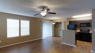 This single-floor space offers a thoughtfully designed open layout that combines comfort and functionality. The main living area features rich hardwood-style flooring that flows throughout, anchored by a ceiling fan with integrated lighting that keeps the space well-lit and comfortable. A large window fitted with white blinds allows natural light to fill the room, while sliding glass doors with vertical blinds provide access to the outdoors and further brighten the interior. A pendant light fixture elegantly marks the transition between the living area and the adjoining dining or kitchen space. The kitchen sits just off the main living area and is partially separated by a raised bar counter finished with granite-style countertops, creating a natural divide while maintaining an open feel. Dark cabinetry overhead adds a bold contrast to the lighter walls, and a built-in appliance niche keeps the space organized. A textured popcorn ceiling runs throughout, adding a classic residential character. The warm, neutral-toned walls tie all elements of the space together, creating a cohesive and inviting ambiance throughout.