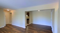 This single-floor residence offers a clean, functional layout designed for comfortable everyday living. The main living area is characterized by rich, dark hardwood flooring that extends throughout, providing a warm and grounded aesthetic against the bright, neutral-toned walls and crisp white baseboards. The space feels open and uncluttered, with a smooth white ceiling and recessed lighting adding a polished finish overhead.

A well-defined opening connects the living area to the kitchen, creating a natural flow between the two spaces while maintaining a sense of visual separation. The kitchen features dark wood cabinetry that extends to the upper walls, providing ample storage. A stainless steel dishwasher is visible beneath the counters, and recessed ceiling lighting illuminates the workspace efficiently. The countertops and backsplash complement the overall dark cabinetry palette.

The overall ambiance of the space is understated and practical, with a neutral color scheme that serves as a versatile backdrop for a variety of interior styles. The consistent flooring and cohesive design language throughout lend the space a unified, well-maintained appearance. The layout and finishes make it a functional and adaptable living environment.