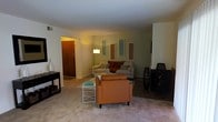 This single-floor residence centers around a welcoming living room that sets a warm, relaxed ambiance through its soft, ambient lighting and neutral color palette. The living area is carpeted and comfortably sized, accommodating a sectional sofa accented with decorative throw pillows and a rust-orange accent chair that adds a pop of warm color to the space. A glass-top coffee table and a stylish side table contribute functional elegance to the seating arrangement.

The walls are painted in a soft sage green tone and are adorned with a series of decorative rectangular wall panels in muted tones of blue, red, and tan, adding visual interest and a contemporary touch to the room. A tall floor lamp provides warm, focused illumination, complementing the overall cozy atmosphere.

A visible entryway with a wooden door is positioned to one side of the living area, offering a clear sense of the layout and flow of the space. Tall decorative vase-like accents and a wall-mounted television round out the room's furnishings, making it both aesthetically pleasing and functionally arranged for everyday living.