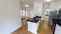 This single-story space showcases an open-concept layout that seamlessly connects the kitchen and living areas, creating a bright and airy atmosphere. The kitchen is anchored by a central island finished with striking dark granite countertops, white cabinetry, and a stainless steel sink. Upper cabinets in crisp white extend toward the ceiling, maximizing storage while maintaining a clean, cohesive aesthetic. Crown molding runs throughout, adding a refined architectural detail to the space.

Warm-toned hardwood flooring flows continuously from the kitchen into the adjacent living area, reinforcing the open feel. The living space benefits from natural light streaming through a set of windows, illuminating the neutral-toned walls and contributing to an inviting ambiance. Flush-mount ceiling fixtures provide additional lighting throughout.

A secondary room, partially visible to the right, appears to feature its own cabinetry and countertop, suggesting a bathroom or utility space. The overall design balances functionality with a clean, modern aesthetic, making efficient use of the available floor plan across its single level.