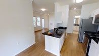This single-story space showcases an open-concept layout that seamlessly connects the kitchen and living areas, creating a bright and airy atmosphere. The kitchen is anchored by a central island finished with striking dark granite countertops, white cabinetry, and a stainless steel sink. Upper cabinets in crisp white extend toward the ceiling, maximizing storage while maintaining a clean, cohesive aesthetic. Crown molding runs throughout, adding a refined architectural detail to the space.

Warm-toned hardwood flooring flows continuously from the kitchen into the adjacent living area, reinforcing the open feel. The living space benefits from natural light streaming through a set of windows, illuminating the neutral-toned walls and contributing to an inviting ambiance. Flush-mount ceiling fixtures provide additional lighting throughout.

A secondary room, partially visible to the right, appears to feature its own cabinetry and countertop, suggesting a bathroom or utility space. The overall design balances functionality with a clean, modern aesthetic, making efficient use of the available floor plan across its single level.