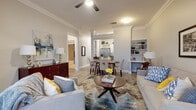 This single-floor residence offers a thoughtfully designed open-concept layout that seamlessly connects the living, dining, and kitchen areas. The living room is anchored by light hardwood flooring, crown molding, a ceiling fan, and built-in shelving, creating an inviting and functional space. A decorative columned archway frames the transition into the dining area, where a six-seat dining table sits beneath additional crown molding detail, with the kitchen visible beyond.

The kitchen features white shaker-style cabinetry, dark granite countertops, and a full suite of stainless steel appliances, including a French door refrigerator, range, microwave, and dishwasher. Light hardwood flooring continues throughout, complementing the clean, bright aesthetic.

A separate sitting room with large windows and French doors offers abundant natural light and a more intimate retreat from the main living area. The bedroom is a serene space with plush carpeting, large windows with natural light, and ample room for a statement four-poster bed. The bathroom includes a soaking tub with shower combination set on tile flooring, providing a calm and practical en-suite environment. The overall ambiance of the space is warm, polished, and move-in ready.