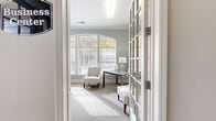 This single-floor space presents a thoughtfully designed office environment that balances functionality with refined aesthetic detail. The main office area is defined by a striking arched window arrangement that floods the room with natural light, framing views of lush outdoor greenery. Soft sage-blue walls complement the neutral carpet flooring and white crown molding, lending the space a calm, professional character.

The office is equipped with a built-in white cabinetry and shelving system that spans an entire wall, providing generous storage and integrated workspace for productivity. A secondary seating area features a striped accent chair and a writing desk, creating a comfortable space for consultation or focused work. Decorative touches, including a large skeletal wall clock and curated shelf displays, add personality without disrupting the clean, cohesive design.

Glass-paned French doors serve as a prominent architectural feature throughout, enhancing the sense of openness while maintaining defined room boundaries. A tiled entryway transitions into the carpeted interior, clearly delineating the entrance from the main work areas. Adjacent spaces, including a pantry and support areas visible through the French doors, reflect thoughtful spatial planning. Overall, the space conveys a welcoming, organized, and polished professional environment.