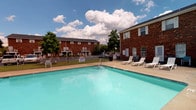 This community space centers around a generously sized outdoor swimming pool with clear, blue-green water, framed by a concrete pool deck equipped with multiple white lounge chairs — ideal for relaxing in the sun. The pool area is enclosed by a chain-link fence and is flanked by classic two-story brick residential buildings that lend a timeless, traditional character to the surroundings. Mature trees and well-kept greenery provide a pleasant backdrop, while open sky above allows for abundant natural light throughout the day. The setting has a clean, functional layout well-suited for outdoor recreation and leisure.

The interior spaces associated with this property feature carpeted rooms finished in neutral, light gray tones with clean white trim and baseboards. The rooms are bright and open, benefiting from a simple, understated design that provides a versatile canvas for a variety of uses. Flush floor vents indicate a central HVAC system, ensuring year-round comfort. The straightforward layout and neutral palette of the interior spaces complement the community-oriented outdoor amenities, offering a cohesive and practical living environment overall.