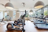 Located at 7400 Parkwood Ct in Falls Church, Virginia, this fitness center offers a generously proportioned, single-floor exercise space designed to support a full range of workout routines. The facility is characterized by its open, airy layout, enhanced by large floor-to-ceiling windows along multiple walls that flood the interior with natural light and provide pleasant outdoor views of the surrounding greenery.

The design features a striking combination of bold red accent walls contrasted against crisp white ceilings, creating an energetic and motivating atmosphere. Distinctive oversized dome pendant lights hang from the ceiling, contributing both to the modern aesthetic and to the bright, evenly lit environment. Durable cork-style flooring runs throughout, offering comfort underfoot.

The cardio zone is well-stocked with multiple treadmills, elliptical trainers, and stationary bikes, all positioned to take advantage of the expansive window views. The strength-training area includes cable and pulley machines, adjustable benches, and a dedicated functional training station. Built-in wooden cubbies along one wall provide convenient storage for accessories and equipment. Full-length mirrors in the strength zone allow users to monitor their form. The space is thoughtfully organized to ensure ample room between stations, supporting a comfortable and safe workout experience.