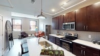 This single-floor urban residence showcases a thoughtfully designed open-concept layout that seamlessly integrates the kitchen and living areas. The kitchen is a standout feature, equipped with rich dark-wood cabinetry extending to the ceiling for ample storage, white quartz countertops, a large island with an undermount sink, and a full suite of stainless steel appliances including a refrigerator, electric range, and over-the-range microwave. A decorative pendant light anchors the island, while exposed ceiling tracks add an industrial character. The living area flows naturally from the kitchen, featuring wood-look flooring, a large window that draws in natural light, and a white-planked ceiling that brightens the space. The bedroom offers a cozy retreat with plush carpeting, soft neutral tones, an exposed whitewashed brick accent wall, and dual windows framing urban cityscape views. The bathroom features wood-look flooring and a clean, understated design. Throughout the unit, industrial-style elements—including exposed brick walls and structural ceiling details—are balanced with modern finishes, creating a cohesive urban aesthetic.