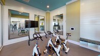 This fitness center is thoughtfully designed to support a comprehensive workout experience across two distinct zones. The space is immediately distinguished by its bold teal ceiling paired with soft olive-green walls, creating an energetic yet focused atmosphere. Durable flooring runs throughout, providing a stable surface suited for a variety of exercise activities.

The cycling area is outfitted with a row of True-brand stationary spin bikes, accompanied by wall-mounted flat-screen televisions and large framed mirrors that amplify the sense of space. A smart control panel is integrated into the wall, offering convenient management of the room's systems.

Adjacent to the cycling zone, the strength and functional training area is equipped with a cable machine, a full dumbbell rack, stability balls in multiple sizes, resistance bands, and additional free weights. Floor-to-ceiling glass panel partitions divide the two areas while maintaining an open, connected feel and allowing natural light to flow freely from large windows.

Strategic placement of mirrors throughout both zones enhances visibility and supports proper form during exercise. Recessed lighting complements the natural light, ensuring the space remains bright and well-illuminated. The overall layout is efficient and welcoming, designed to accommodate a range of fitness activities in a clean, modern environment.