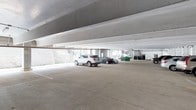 This single-level covered parking facility provides a functional and well-organized space designed to accommodate multiple vehicles with ease. The garage features a flat concrete floor with clearly painted stall markings, ensuring efficient use of the available area and straightforward navigation for drivers.

The structure is supported by exposed concrete beams running across the ceiling, giving the space an industrial yet clean aesthetic. The open-plan layout allows for generous clearance height, accommodating a variety of vehicle types including SUVs and sedans. Natural light filters in from the open perimeter edges of the structure, providing a sense of openness while maintaining the protected environment of a covered garage.

A stairwell is visible toward the rear of the facility, suggesting connectivity to adjacent or upper-level areas of the broader building complex. Utility elements such as waste receptacles are discreetly positioned near the stairwell access point. The concrete construction throughout — floors, columns, and ceiling — conveys a durable, low-maintenance design built for long-term use. Overall, this parking facility offers a practical, accessible, and well-lit environment suited to serve a residential, commercial, or mixed-use property.