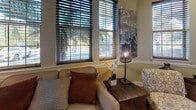 This single-floor space combines a warm, professional atmosphere with thoughtful design elements throughout. The main lounge area is a standout feature, boasting soaring vaulted ceilings with an arched transom window that floods the room with natural light. A bank of large windows dressed with wood-toned blinds offers views of lush outdoor greenery, including palm trees, creating a bright and inviting ambiance. Rich hardwood flooring flows seamlessly across the entire space, anchoring the warm neutral color palette of soft yellows and taupes accented by crisp white crown molding and wainscoting. The lounge is furnished with a comfortable sofa and accent chairs, making it an ideal area for relaxed meetings or waiting. Adjacent to the lounge is a private office area, enclosed by elegant French doors with multi-pane glass inserts that maintain an open feel while providing separation. The office is equipped with a dark wood desk, ergonomic seating, an open shelving unit, and its own window for natural light. Safety and accessibility features, including emergency signage and security hardware, are discreetly integrated. Overall, the space strikes a balance between professional functionality and a welcoming, homey character.
