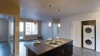This single-floor space showcases an open-concept layout that integrates the kitchen and living areas into one cohesive, functional environment. The kitchen island serves as the focal point, featuring dark cabinetry paired with rich, dark granite countertops and a double stainless steel sink with a classic-style faucet. Three cylindrical pendant lights hang at varying heights above the island, providing both task lighting and a stylish design element with their warm, frosted glass shades.

The dark lower cabinetry of the island contrasts effectively against the neutral grey walls and light-toned ceiling, creating a balanced visual appeal throughout. The flooring is a smooth, dark surface that runs consistently through both the kitchen and the adjacent living area, reinforcing the open, unified feel of the space.

The living area beyond the island benefits from generous natural light streaming through a large window and sliding glass door, illuminating the room and offering views of the surrounding exterior. A paneled interior door on the opposing wall provides access to additional areas of the home. The overall ambiance is modern and clean, combining functional design with contemporary finishes.