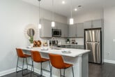 This thoughtfully designed one-bedroom, one-bathroom unit offers a modern, open-concept layout that maximizes every inch of its compact footprint. The living and kitchen areas flow seamlessly together, anchored by rich, dark hardwood-style flooring and a neutral gray-and-white color palette that lends a clean, contemporary feel throughout.

The kitchen is a standout feature, equipped with gray shaker-style cabinetry, a white quartz countertop, and a full suite of stainless steel appliances including a refrigerator, range, microwave, and dishwasher. A white subway tile backsplash adds a classic touch, while three pendant lights hang above the peninsula breakfast bar, which comfortably seats three and serves as both a dining and entertaining space.

The bedroom is a quiet retreat, featuring plush carpeting, soft neutral walls, and a generous window that allows natural light to filter in. The bathroom maintains the same cohesive design language, with gray cabinetry, a white vanity top, and light tile flooring. A laundry area is also incorporated within the unit for added convenience. Recessed lighting and tall ceilings contribute to an airy, open ambiance throughout the space.