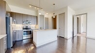 This single-floor unit near SH-99 in Houston's Katy area showcases a contemporary design with a neutral, light-filled palette throughout. The open-concept kitchen serves as a functional centerpiece, featuring dark wood-tone cabinetry, a full suite of stainless steel appliances including a refrigerator, range, microwave, and dishwasher, and a white kitchen island with an integrated sink and pendant lighting overhead. Track lighting runs along the elevated ceiling, adding both task and ambient illumination. Rich, dark wood-style flooring extends through the kitchen and main living areas, complementing the clean white walls and modern millwork. The living area is flooded with natural light through an expansive floor-to-ceiling window arrangement, with transom windows above offering additional sky views and outdoor greenery. A separate room features built-in cabinetry and a desk, offering functional workspace potential. Bedrooms are finished with plush carpeting and large windows. The bathroom includes a full tub and tile surround, a wide vanity with quartz countertops, dark wood cabinetry, a large mirror, and wood-style flooring, completing the cohesive modern aesthetic found throughout the unit.