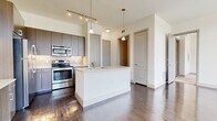 This single-floor unit near SH-99 in Houston's Katy area showcases a contemporary design with a neutral, light-filled palette throughout. The open-concept kitchen serves as a functional centerpiece, featuring dark wood-tone cabinetry, a full suite of stainless steel appliances including a refrigerator, range, microwave, and dishwasher, and a white kitchen island with an integrated sink and pendant lighting overhead. Track lighting runs along the elevated ceiling, adding both task and ambient illumination. Rich, dark wood-style flooring extends through the kitchen and main living areas, complementing the clean white walls and modern millwork. The living area is flooded with natural light through an expansive floor-to-ceiling window arrangement, with transom windows above offering additional sky views and outdoor greenery. A separate room features built-in cabinetry and a desk, offering functional workspace potential. Bedrooms are finished with plush carpeting and large windows. The bathroom includes a full tub and tile surround, a wide vanity with quartz countertops, dark wood cabinetry, a large mirror, and wood-style flooring, completing the cohesive modern aesthetic found throughout the unit.
