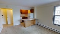 This single-floor unit is designed with a practical, open layout that connects the kitchen and living area seamlessly. The kitchen is equipped with warm-toned wood cabinetry mounted above and below the counter, providing ample storage space. Appliances include a white refrigerator, a gas range, and a sink positioned along the back wall beneath the upper cabinets. A peninsula counter with light wood cabinetry extends outward from the kitchen, creating a natural dividing point between the cooking and living spaces while also offering additional prep or dining surface area. The transition from the kitchen's hard flooring to the carpeted living area is clearly defined, adding a sense of distinction between functional zones. The living space is generously sized and open, with neutral-toned carpet and plain white walls that provide a clean, versatile backdrop. A baseboard heater runs along the perimeter wall of the living area, indicating an electric heating system. The overall aesthetic is simple and functional, with soft, warm tones from the cabinetry complementing the neutral palette of the walls and flooring throughout the space.