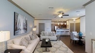 This single-floor residence showcases an open-concept design that seamlessly integrates the living, dining, and kitchen areas into one cohesive, flowing space. The living room is anchored by a ceiling fan with a light fixture and features soft blue-gray accent walls adorned with large-scale artwork, creating a sophisticated yet welcoming ambiance. Cream-toned sofas and neutral armchairs are arranged around a patterned area rug, complementing the room's calm, neutral palette.

The dining area incorporates a glass-topped table surrounded by upholstered chairs, offering an elegant space for meals. Multiple windows dressed with navy blue curtains and horizontal blinds allow generous natural light to filter throughout the space.

The kitchen, visible from the living area, features dark wood cabinetry, stainless steel appliances, pendant lighting, and a breakfast bar with bar stools, providing both functionality and style. Dark hardwood-style flooring in the kitchen transitions smoothly to plush carpeting in the living area. Crown molding details throughout the space add a refined architectural touch, contributing to the overall polished and inviting atmosphere.