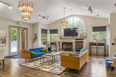This residential lounge is a generously sized, single-floor community space designed to serve multiple functions within a cohesive, modern aesthetic. The layout is divided into distinct yet open zones, each thoughtfully furnished and decorated.

The living area is the centerpiece of the lounge, featuring vaulted ceilings that amplify the sense of openness. A bold blue velvet sofa anchored on a geometric black-and-white area rug sets a mid-century modern tone, complemented by warm wood accents, mustard yellow throw pillows, and layered artwork in navy and white. A tiered gold chandelier and track lighting illuminate the space, while multiple windows with navy roller shades invite natural light and views of surrounding greenery. A fireplace with built-in shelving adds a residential warmth to the room.

Adjacent to the living area is a casual dining and kitchen nook, equipped with counter seating, a small kitchen pass-through, and direct access to an outdoor patio through glass doors.

A business center occupies a separate wing, featuring vaulted skylights, structural white columns, wood-look flooring, and individual workstations with computers—ideal for residents seeking a productive environment. The overall ambiance throughout is bright, modern, and welcoming.