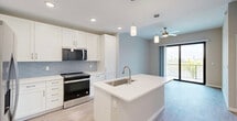 Located in Bonita Springs, FL, this well-designed one-bedroom, one-bathroom unit offers a clean, modern aesthetic throughout. The open-concept layout seamlessly connects the kitchen, dining, and living areas, creating a comfortable and functional living space. The kitchen is a standout feature, boasting crisp white shaker cabinetry, a light blue tile backsplash, stainless steel appliances including a refrigerator with ice maker, electric range, built-in microwave, and dishwasher, all centered around a large white quartz island with an undermount sink and pendant lighting above. Light wood-style flooring runs through the main living areas, complementing the soft, neutral color palette. The living area is illuminated by a ceiling fan and opens directly to a private patio through sliding glass doors, offering outdoor views and natural light. The spacious bedroom features plush carpeting, a ceiling fan, and a generously sized window. The bathroom is thoughtfully appointed with a wide single-sink vanity with white cabinetry, a large mirror, a tub-shower combination, and access to a walk-in closet with built-in shelving. Two closets provide ample storage throughout the unit. The overall ambiance is bright, clean, and contemporary.