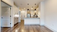 This 540 sq ft, one-bedroom, one-bathroom apartment at 4177 Gazebo Street in Aubrey, TX features an open-concept kitchen and living area with wood-look flooring throughout. The kitchen is equipped with stainless steel appliances, granite countertops, a large island with pendant lighting, white upper cabinets, warm wood lower cabinets, and a herringbone tile backsplash. The 120 sq ft bedroom offers natural light through multiple windows with outdoor views. The bathroom includes a granite-topped vanity and direct access to a walk-in closet with built-in shelving.