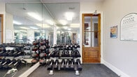 This fitness center is a comprehensively outfitted exercise facility designed to support a full range of workout routines. The space is accessed through a charming wood-framed glass-panel door and is finished with durable speckled rubber flooring, bright recessed panel lighting, and clean white walls that create a crisp, energizing atmosphere.

Floor-to-ceiling mirrors line multiple walls throughout the facility, visually expanding the space and allowing users to monitor their form during workouts. The cardio section features a lineup of True-brand treadmills, each equipped with individual flat-screen monitors, as well as spin bikes and additional cycle machines. These are positioned to take advantage of natural light streaming in through several windows, which offer views of the surrounding outdoor areas including what appears to be a pool patio.

The free weights area houses a well-organized three-tier dumbbell rack with a broad range of weights, complemented by a dedicated medicine ball rack. Strength training equipment is also visible throughout the space, offering options for resistance and machine-based training.

A posted Fitness Center sign notes that the facility operates 24 hours a day, making it a highly accessible amenity. The combination of traditional architectural details, modern equipment, and thoughtful layout makes this fitness center both functional and welcoming.