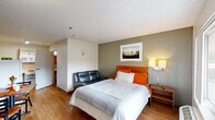 This compact studio unit in Baton Rouge, Louisiana, is thoughtfully designed to maximize functionality within a single open-plan floor. The main living area serves as the bedroom, sitting area, and dining space combined, anchored by a queen-sized bed with a warm wood headboard, orange accent bedding, and a bedside nightstand with a lamp. A small leather sofa sits adjacent to the bed, providing a secondary seating option. A window along one wall allows natural light to filter in, offering a view of the exterior surroundings.

The dining area features a modest table with chairs, accented with orange placemats, and a wall-mounted flat-screen television is positioned nearby for convenient viewing. The kitchenette is tucked along one wall and includes a compact refrigerator, upper cabinetry, and a counter workspace, providing essential food preparation capabilities. Light wood-tone flooring runs throughout the unit, lending warmth and visual continuity to the space.

The private bathroom is outfitted with accessibility features, including stainless steel grab bars positioned near the toilet and along the wall, a pedestal sink, and wood-tone flooring consistent with the main area. The textured ceiling and neutral wall tones complete the unit's straightforward, practical aesthetic.
