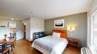 This compact studio unit in Baton Rouge, Louisiana, is thoughtfully designed to maximize functionality within a single open-plan floor. The main living area serves as the bedroom, sitting area, and dining space combined, anchored by a queen-sized bed with a warm wood headboard, orange accent bedding, and a bedside nightstand with a lamp. A small leather sofa sits adjacent to the bed, providing a secondary seating option. A window along one wall allows natural light to filter in, offering a view of the exterior surroundings.

The dining area features a modest table with chairs, accented with orange placemats, and a wall-mounted flat-screen television is positioned nearby for convenient viewing. The kitchenette is tucked along one wall and includes a compact refrigerator, upper cabinetry, and a counter workspace, providing essential food preparation capabilities. Light wood-tone flooring runs throughout the unit, lending warmth and visual continuity to the space.

The private bathroom is outfitted with accessibility features, including stainless steel grab bars positioned near the toilet and along the wall, a pedestal sink, and wood-tone flooring consistent with the main area. The textured ceiling and neutral wall tones complete the unit's straightforward, practical aesthetic.