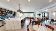 Located at 17120 North 53rd Avenue in Glendale, Arizona, this single-story community clubhouse offers a generously proportioned, thoughtfully designed gathering space. The open-concept layout connects a fully equipped kitchen to multiple dining and lounge areas, creating a seamless flow throughout.

The kitchen is a standout feature, anchored by a sweeping marble-look island with gold veining, complemented by white shaker cabinetry with matte black hardware and a bold navy-and-gold chevron tile backsplash. Woven rattan pendant lights add a warm, textural contrast to the crisp white finishes. Stainless steel appliances and open metal shelving complete the functional yet stylish setup.

The dining area accommodates a variety of seating arrangements, from communal long tables to intimate round bistro tables, all set across patterned tilework and light hardwood-style flooring. Lush greenery displayed on overhead shelving brings a fresh, natural element to the space, while large windows and glass doors invite abundant natural light and views of the outdoor surroundings.

A dedicated game room provides a lively entertainment zone, featuring a full-size pool table, a round poker table, a wall-mounted television, and a patterned area rug beneath a modern linear chandelier. Adjacent lounge seating with built-in shelving and a decorative accent wall rounds out this versatile, amenity-rich space.