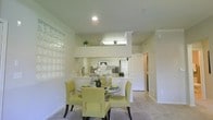 This single-floor residence offers a thoughtfully designed open-concept layout that seamlessly connects the dining and kitchen areas, creating an inviting and functional living environment. The dining area is a standout space, highlighted by a striking wall of glass block windows that diffuse soft, natural light throughout the room, lending a warm and airy ambiance. The space comfortably accommodates a round glass-top dining table with elegant chartreuse upholstered chairs, adding a pop of color against the neutral palette.

The kitchen is positioned adjacent to the dining area, separated by a raised breakfast bar that doubles as a serving counter. Crisp white cabinetry, a white refrigerator, and a microwave above the range contribute to a clean, cohesive aesthetic. A mirrored backsplash behind the kitchen adds depth and brightness to the space. Decorative greenery and vases atop the upper kitchen partition add a welcoming, lived-in touch.

The interiors feature crown molding, recessed lighting, and neutral carpeting that run throughout, contributing to a polished and cohesive finish. A hallway visible beyond the kitchen suggests additional rooms, reinforcing the practical and well-organized floor plan. Overall, the space balances style and functionality in a bright, well-appointed setting.