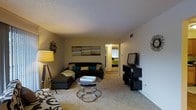 This single-story residence features a welcoming living room as its central gathering space. The room is finished with wall-to-wall carpet in a neutral tone, contributing to a comfortable and cohesive atmosphere. A textured popcorn ceiling adds a classic architectural detail overhead, while a ceiling vent indicates a central HVAC system throughout.

The living room is laid out to accommodate a sectional sofa accented with teal and patterned throw pillows, complemented by a matching ottoman. A set of nesting coffee tables with chrome frames serves as a functional and stylish centerpiece. Entertainment needs are addressed by a multi-tiered media console positioned along one wall, supporting a flat-screen television.

Warm ambient lighting from a floor lamp enhances the room's inviting tone. Wall décor, including a multi-panel canvas piece, adds visual interest to the neutral walls. A hallway visible from the living area leads to a bedroom, suggesting a practical and connected floor plan. The overall aesthetic is modern and functional, with a clean, organized layout designed for everyday living.