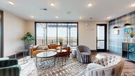 Located at 2010 N Brevard St in Charlotte's NoDa neighborhood, this single-floor amenity lounge offers a thoughtfully designed communal retreat with a seamless blend of indoor comfort and outdoor access. The main lounge area is anchored by a large seating arrangement featuring a leather sofa, tufted accent chairs, and layered area rugs atop warm wood-look flooring. Floor-to-ceiling windows with sleek black frames flood the space with natural light and frame expansive views of the surrounding Charlotte skyline and urban landscape.

A full wet bar runs along one wall, complete with dark navy cabinetry, a white countertop, a built-in beverage cooler, and a mounted large-screen television above — ideal for gatherings and entertaining. Decorative perforated metal panels and white subway tile accents add an industrial-chic character to this area.

An adjoining lounge nook features a striking plaid-patterned accent wall adorned with framed artwork and bowler hat wall sconces, providing a whimsical, curated aesthetic. A storage credenza with display shelving completes the look.

Direct access leads to an open-air balcony with a pergola structure overhead, metal railings, and seating that overlooks the surrounding residential community — offering a relaxing outdoor escape with commanding elevated views of Charlotte.