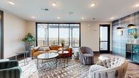 Located at 2010 N Brevard St in Charlotte's NoDa neighborhood, this single-floor amenity lounge offers a thoughtfully designed communal retreat with a seamless blend of indoor comfort and outdoor access. The main lounge area is anchored by a large seating arrangement featuring a leather sofa, tufted accent chairs, and layered area rugs atop warm wood-look flooring. Floor-to-ceiling windows with sleek black frames flood the space with natural light and frame expansive views of the surrounding Charlotte skyline and urban landscape.

A full wet bar runs along one wall, complete with dark navy cabinetry, a white countertop, a built-in beverage cooler, and a mounted large-screen television above — ideal for gatherings and entertaining. Decorative perforated metal panels and white subway tile accents add an industrial-chic character to this area.

An adjoining lounge nook features a striking plaid-patterned accent wall adorned with framed artwork and bowler hat wall sconces, providing a whimsical, curated aesthetic. A storage credenza with display shelving completes the look.

Direct access leads to an open-air balcony with a pergola structure overhead, metal railings, and seating that overlooks the surrounding residential community — offering a relaxing outdoor escape with commanding elevated views of Charlotte.