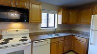 This single-floor space includes a kitchen designed for practical everyday use. The kitchen features an L-shaped layout that maximizes counter space, finished with granite-style countertops in earthy brown and gray tones. Warm medium-toned oak cabinetry lines both the upper and lower walls, providing ample storage throughout. White appliances, including a coil-burner electric range, dishwasher, and an over-the-counter black microwave, lend a clean, classic appearance. A stainless steel sink is centrally positioned beneath a window framed in black, which overlooks an adjacent brick exterior, allowing natural light to filter into the space. The lower cabinets include a combination of door-and-drawer configurations, offering versatile storage options. The countertops extend along two walls in an efficient wraparound design, providing generous prep space. The overall aesthetic is straightforward and functional, with neutral wall tones complementing the wood cabinetry and stone-look surfaces, creating a cohesive and practical kitchen environment suited for daily use.