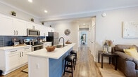 This well-appointed single-floor residence showcases a thoughtfully designed open-concept layout that seamlessly connects the kitchen, dining, and living areas. The kitchen is a standout feature, boasting white shaker cabinetry, a striking blue shiplap island with a built-in sink and bar seating for two, stainless steel appliances, and a navy blue tile backsplash that adds a bold contrast to the crisp white countertops. Recessed lighting and crown molding throughout lend a polished, cohesive aesthetic to the main living spaces. The living room offers comfortable proportions, a wall-mounted TV, and access to a private exterior door, filling the space with natural light. A separate dining nook provides an intimate setting for meals. The primary bedroom is generously sized, featuring multiple windows dressed with neutral linen curtains that invite abundant natural light, and connects to a well-appointed bathroom with white shaker vanity cabinetry, black hardware accents, and hardwood-style flooring. A second bedroom mirrors the serene, light-filled character of the primary suite. Warm hardwood-style flooring flows consistently throughout the entire residence, unifying the space with a modern yet inviting character.